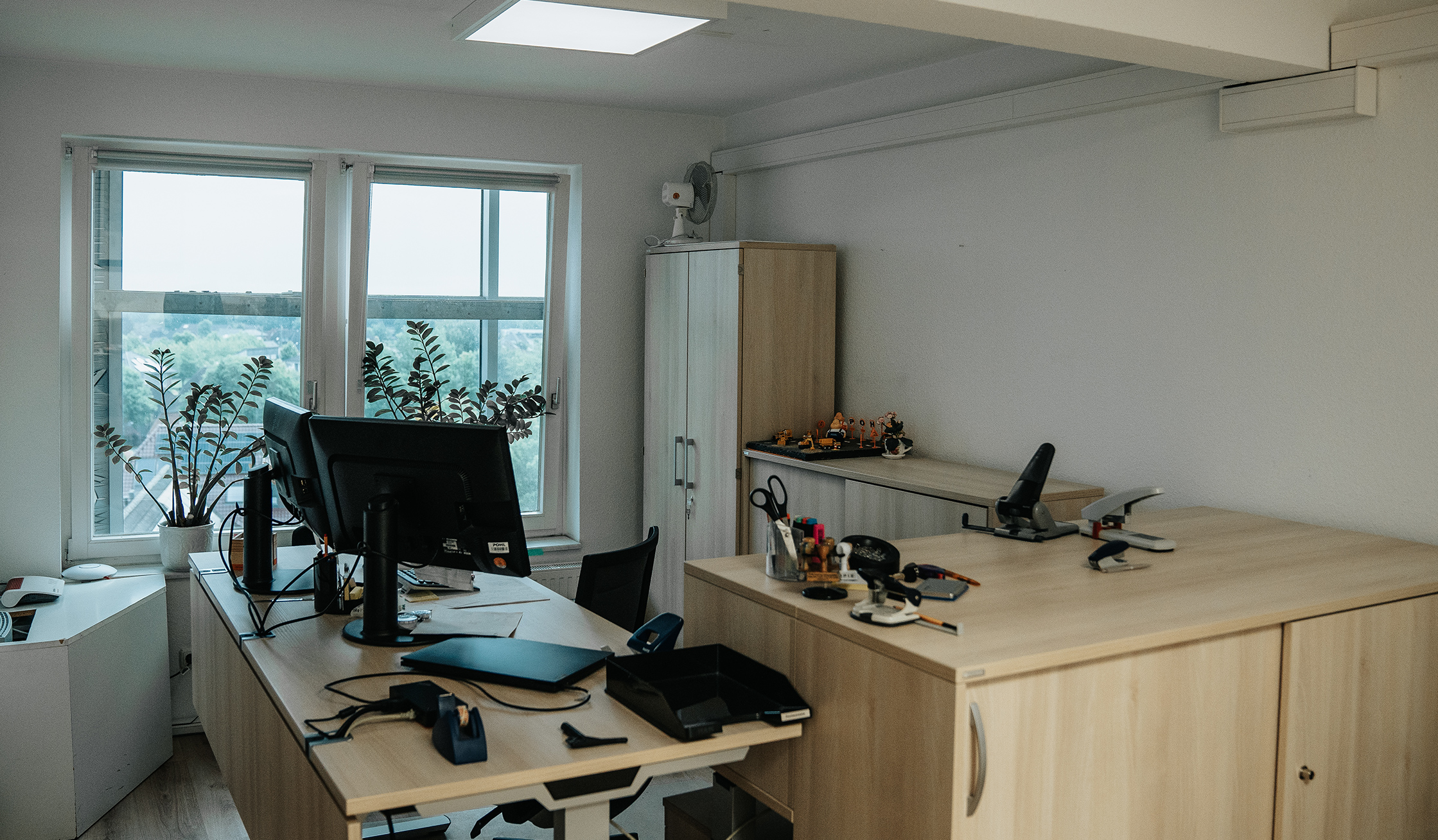 Standort Bönningstedt: Vorne im Bild steht ein Schreibtisch mit zwei Monitoren und Büro-Utensilien. In der Mitte fällt Tageslicht durch zwei Fenster; davor stehen Pflanzen in weißen Töpfen. Rechts an der Wand steht ein hoher Schrank. Im Hintergrund ist über die Fenster eine Stadtlandschaft zu sehen.