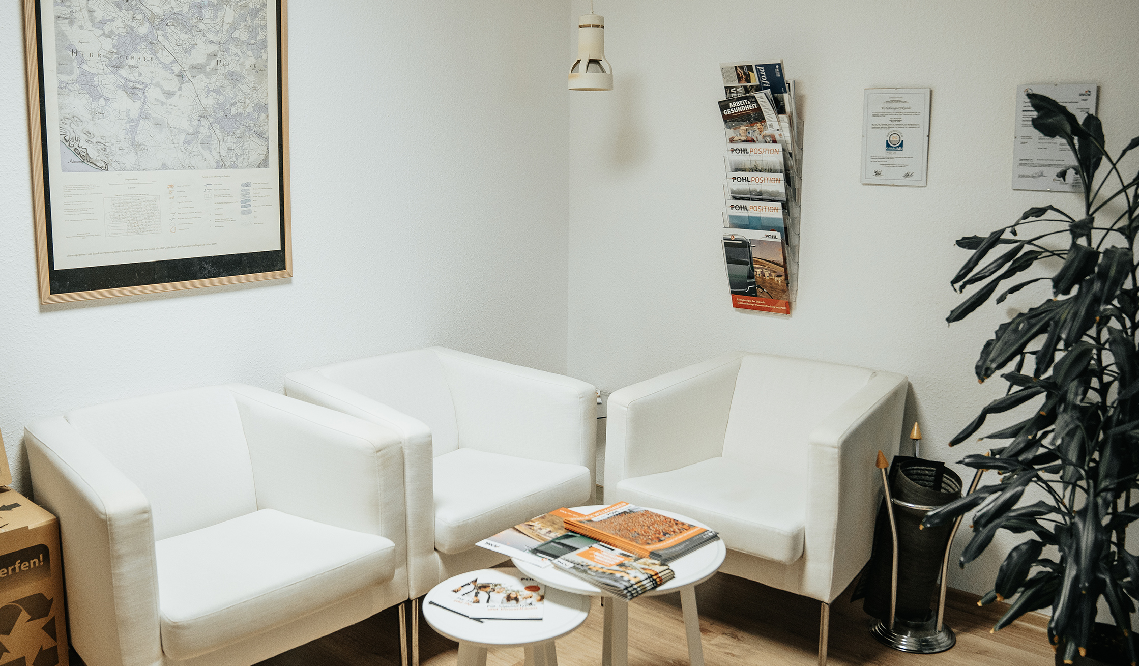 Standort Bönningstedt: Vorne im Bild stehen drei weiße Sessel um zwei kleine runde Tische. In der Mitte liegen auf den Tischen Broschüren, darunter Hefte mit POHL-Aufdruck. Rechts an der Wand hängt ein Prospekthalter mit weiteren POHL-Broschüren. Links im Bild hängt eine große Karte in einem Rahmen.