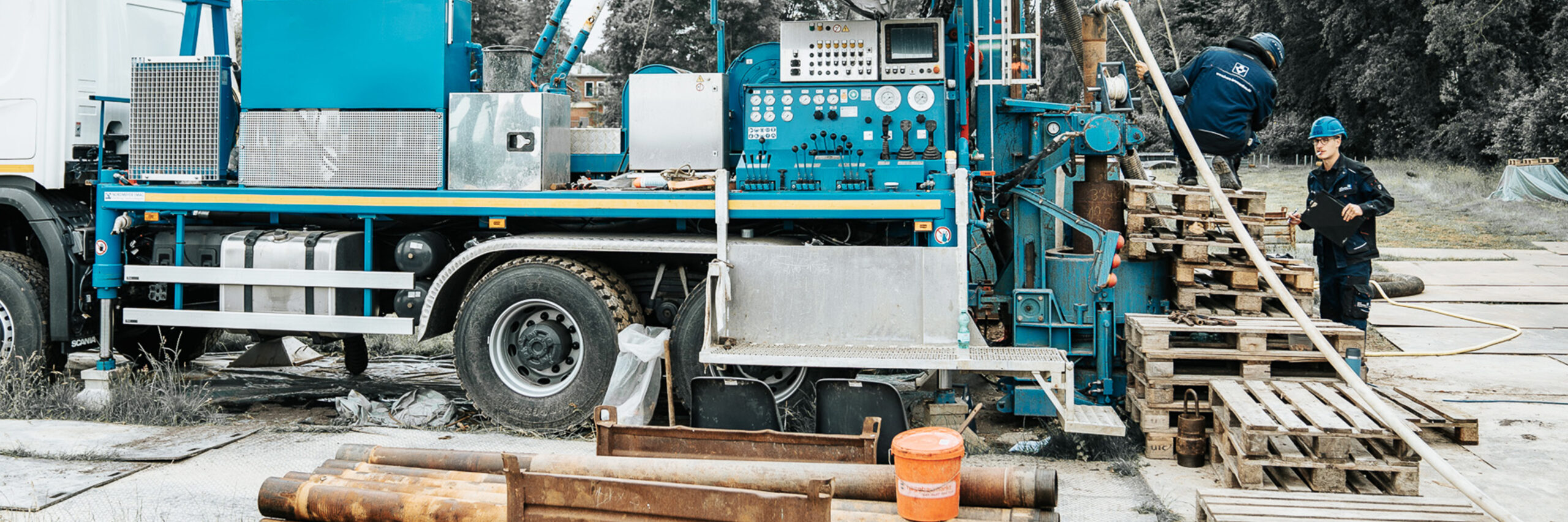 Auf einer Baustelle ist mittig ein großes blaues Bohrgerät auf einem LKW montiert. Links im Bild erkennt man das Fahrerhaus des Lastwagens, teilweise abgeschnitten. Im unteren Vordergrund liegen mehrere Metallrohre und ein orangefarbener Eimer. In der Mitte dominiert das Bohrgerät mit zahlreichen Bedienhebeln, Anzeigen und Schläuchen. Rechts im Bild stapeln sich Holzpaletten, auf denen zwei Arbeiter tätig sind: Einer kniet oben auf den Paletten und beugt sich nach vorn zur Maschine, ein anderer steht unten rechts, trägt einen blauen Helm und hält einen schwarzen Klemmbrettordner. Der Hintergrund zeigt eine Wiese und Bäume.