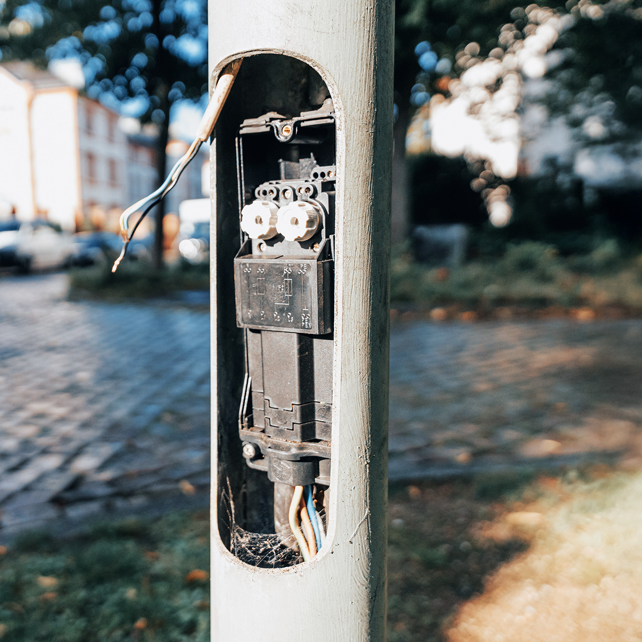 Referenz Wartung Straßenbeleuchtung Büdelsdorf: Vordergrund zeigt eine geöffnete Klappe am Laternenmast Kabel und ein Bauteil. Hinten links stehen Fahrzeuge auf Kopfsteinpflaster, dahinter Häuser unscharf.