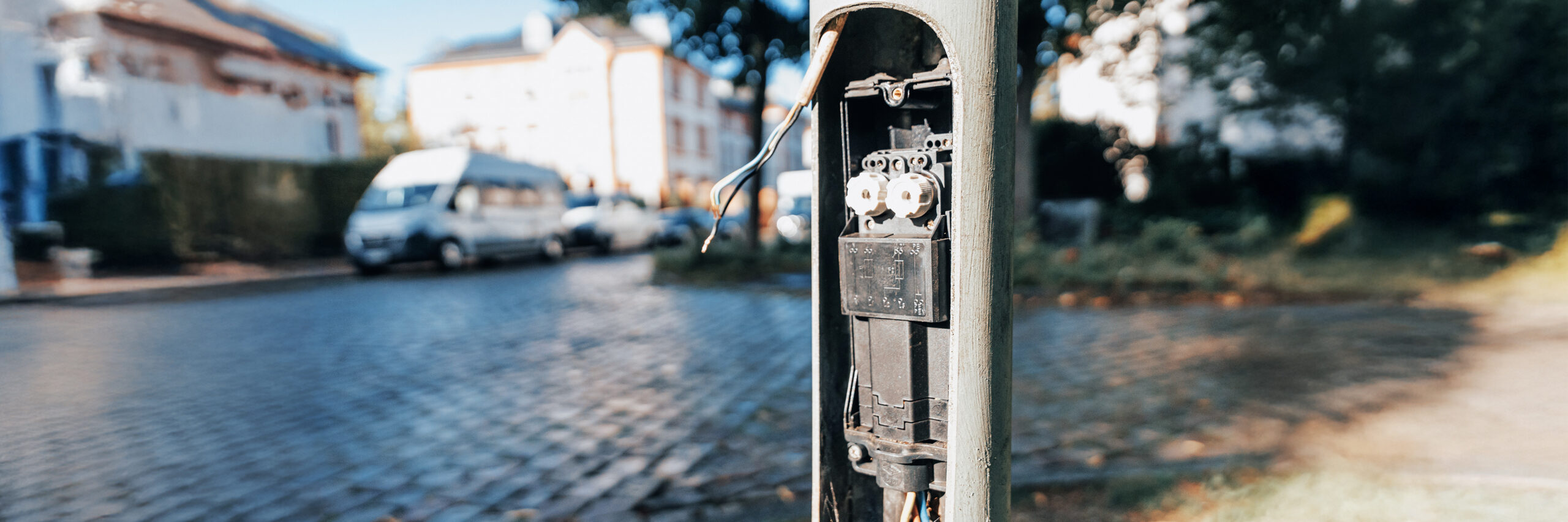 Referenz Wartung Straßenbeleuchtung Büdelsdorf: Vordergrund zeigt eine geöffnete Klappe am Laternenmast Kabel und ein Bauteil. Hinten links stehen Fahrzeuge auf Kopfsteinpflaster, dahinter Häuser unscharf.