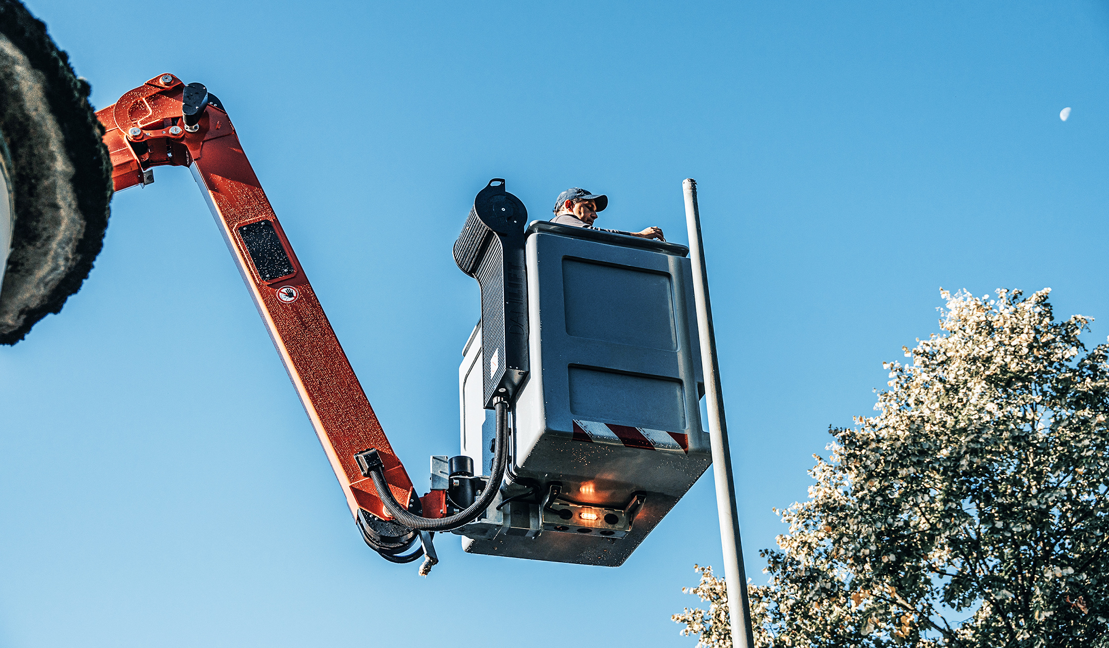 Referenz Wartung Straßenbeleuchtung Büdelsdorf: Unten links hebt der orange Ausleger eines POHL-Fahrzeugs den grauen Arbeitskorb. In der Mitte steht ein POHL-Mitarbeiter neben einem Laternenmast; rechts eine Baumkrone; darüber blauer Himmel.