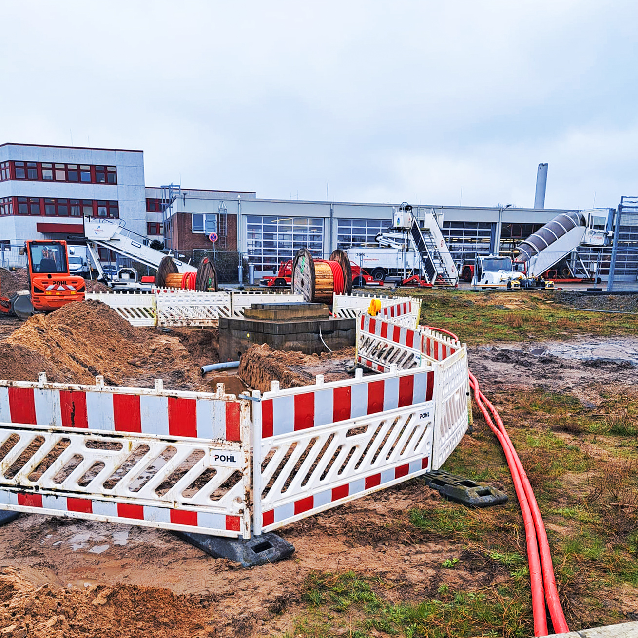 Referenz Stromversorgung Hamburg Flughafen: Vorne im Bild liegt eine abgesperrte Baustelle mit aufgeschüttetem Sand und mehreren großen Kabeltrommeln mit rotem Kabel. Vorne links im Bild steht eine orange POHL-Maschine mit Schaufel. Im Hintergrund erstreckt sich ein langes Hallengebäude mit großen Glastoren; davor stehen weiße Dienstfahrzeuge und zwei mobile Treppen auf Rädern.