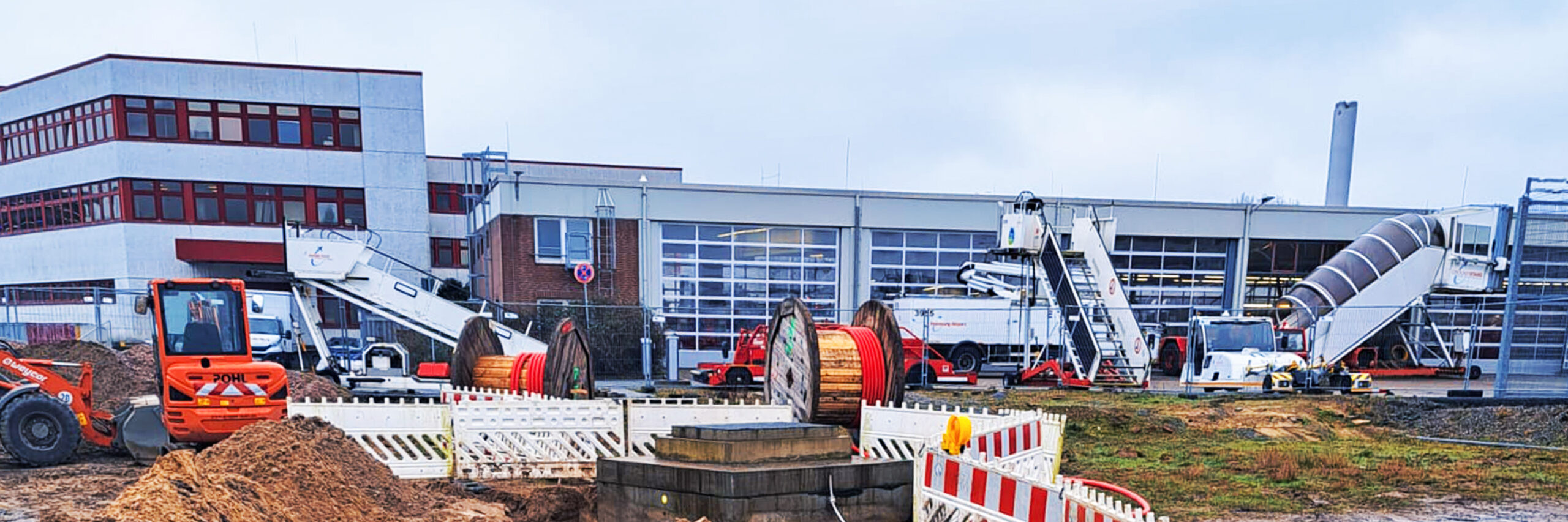 Referenz Stromversorgung Hamburg Flughafen: Vorne im Bild liegt eine abgesperrte Baustelle mit aufgeschüttetem Sand und mehreren großen Kabeltrommeln mit rotem Kabel. Vorne links im Bild steht eine orange POHL-Maschine mit Schaufel. Im Hintergrund erstreckt sich ein langes Hallengebäude mit großen Glastoren; davor stehen weiße Dienstfahrzeuge und zwei mobile Treppen auf Rädern.