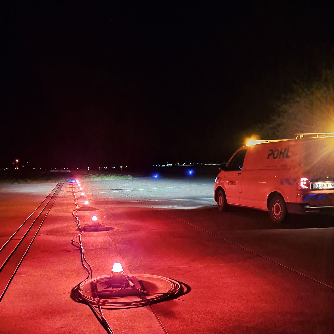 Referenz Landebahnbefeuerung Hamburg Finkenwerder: In der Nacht steht ein orangefarbener Transporter von POHL auf dem Rollfeld. Rundherum leuchten rote und blaue Bodenlichter