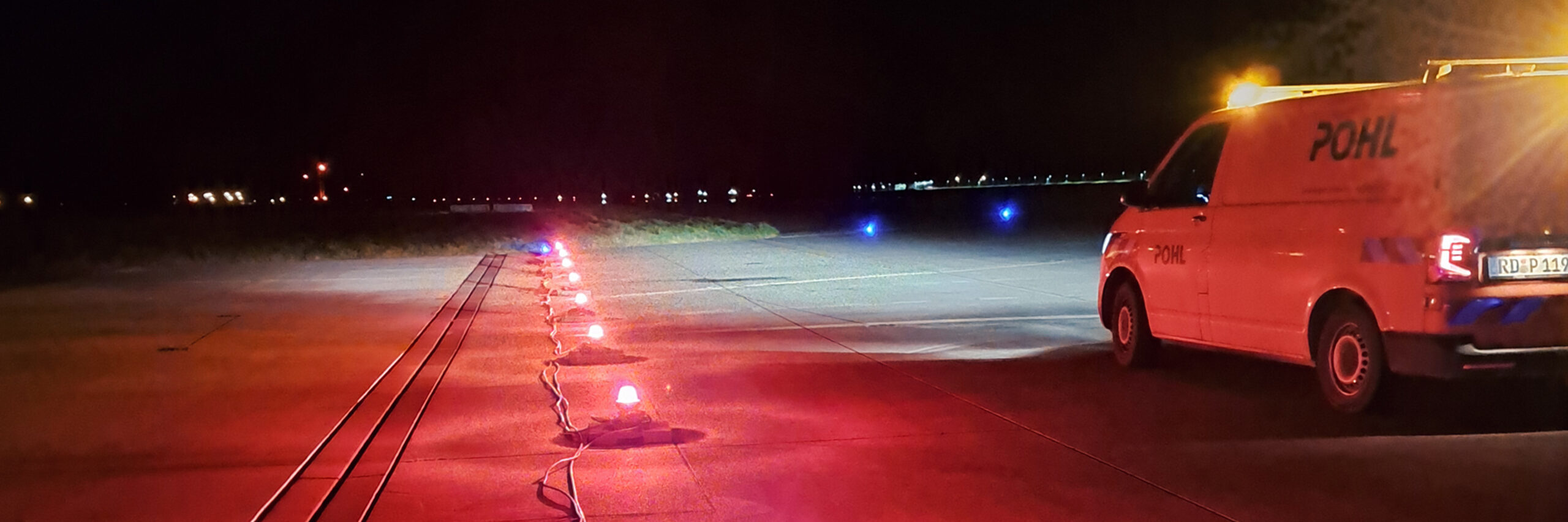 Referenz Hamburg Finkenwerder Landebahnbefeuerung: In der Nacht steht ein orangefarbener Transporter von POHL auf dem Rollfeld. Rundherum leuchten rote und blaue Bodenlichter