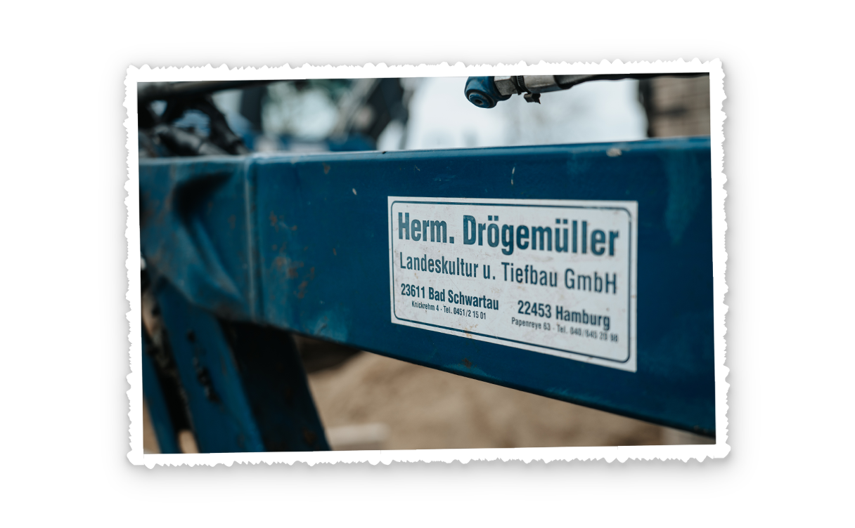 Nahaufnahme eines blauen Maschinenarms mit weißem Herstelleraufkleber, im Vordergrund die abgenutzte Kante, unscharfer Baustellenhintergrund.