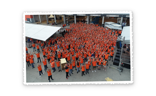 Luftaufnahme einer großen Menschenmenge in orangefarbenen T-Shirts, viele heben die Arme, auf dem Betriebshof sind Zelte und Spielmodule zu sehen, auf den Absperrbannern im Hintergrund steht der Pohl Schriftzug.