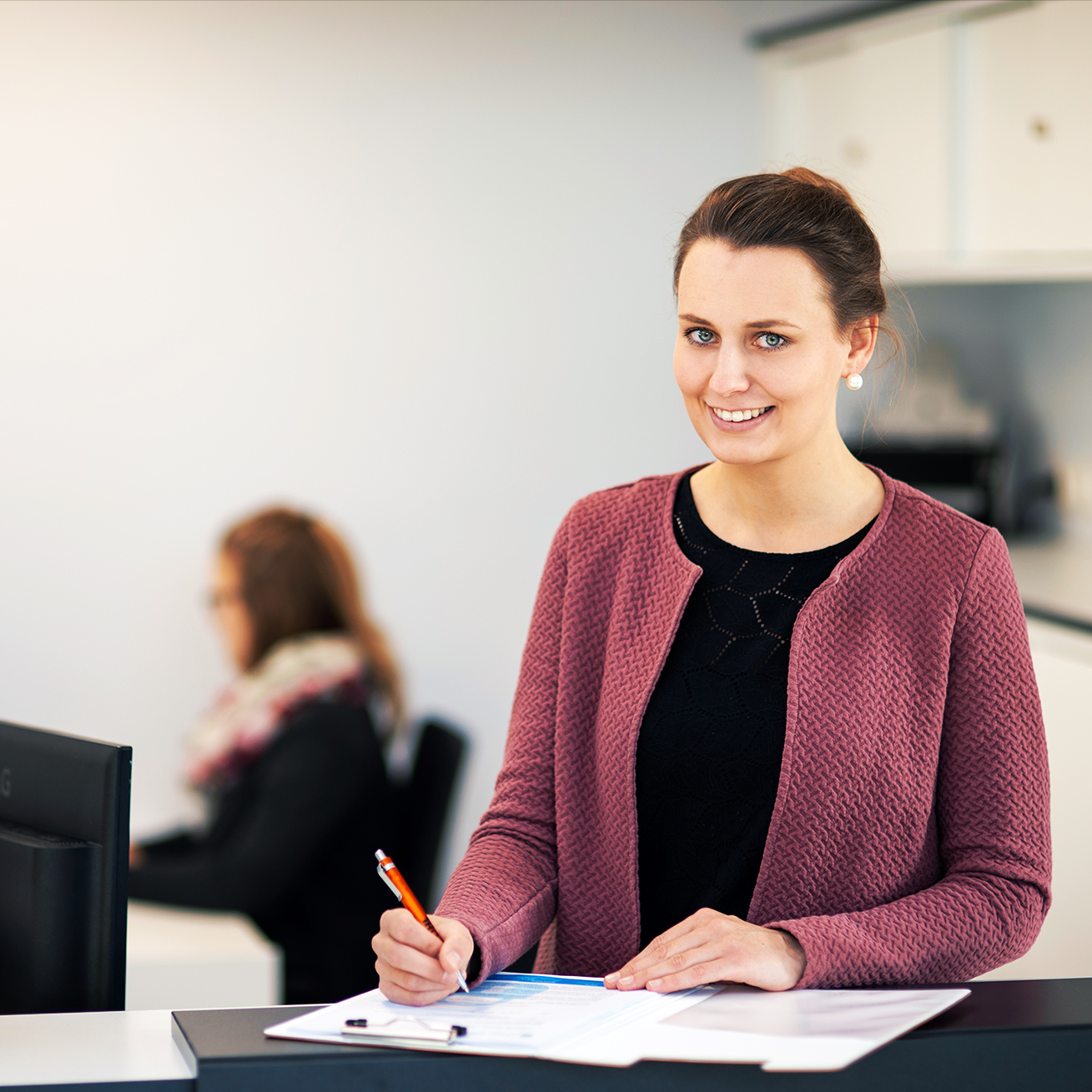 In der Mitte füllt eine POHL-Mitarbeiterin an einem Tresen ein Formular aus. Links vorne steht eine Schale mit Pflanzen; rechts auf der Theke stehen zwei kleine Figuren mit POHL-Aufdruck. Im Hintergrund sitzt eine weitere Person am Schreibtisch.
