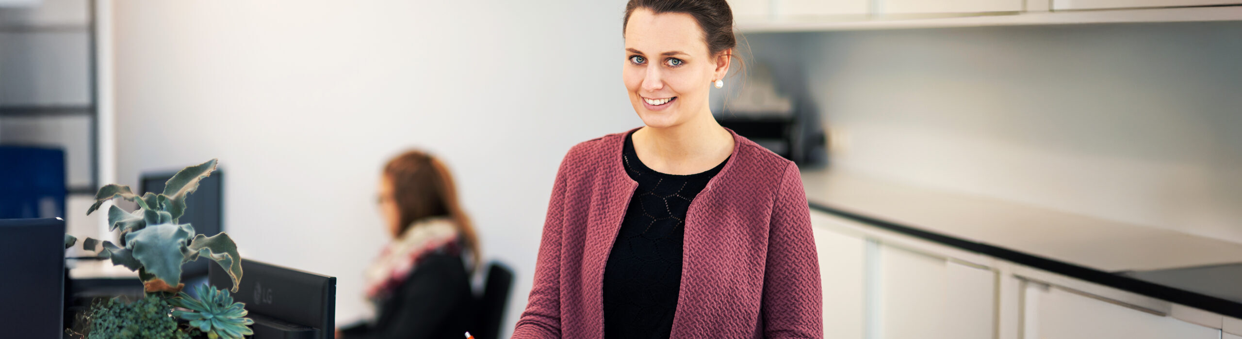 In der Mitte füllt eine POHL-Mitarbeiterin an einem Tresen ein Formular aus. Links vorne steht eine Schale mit Pflanzen; rechts auf der Theke stehen zwei kleine Figuren mit POHL-Aufdruck. Im Hintergrund sitzt eine weitere Person am Schreibtisch.
