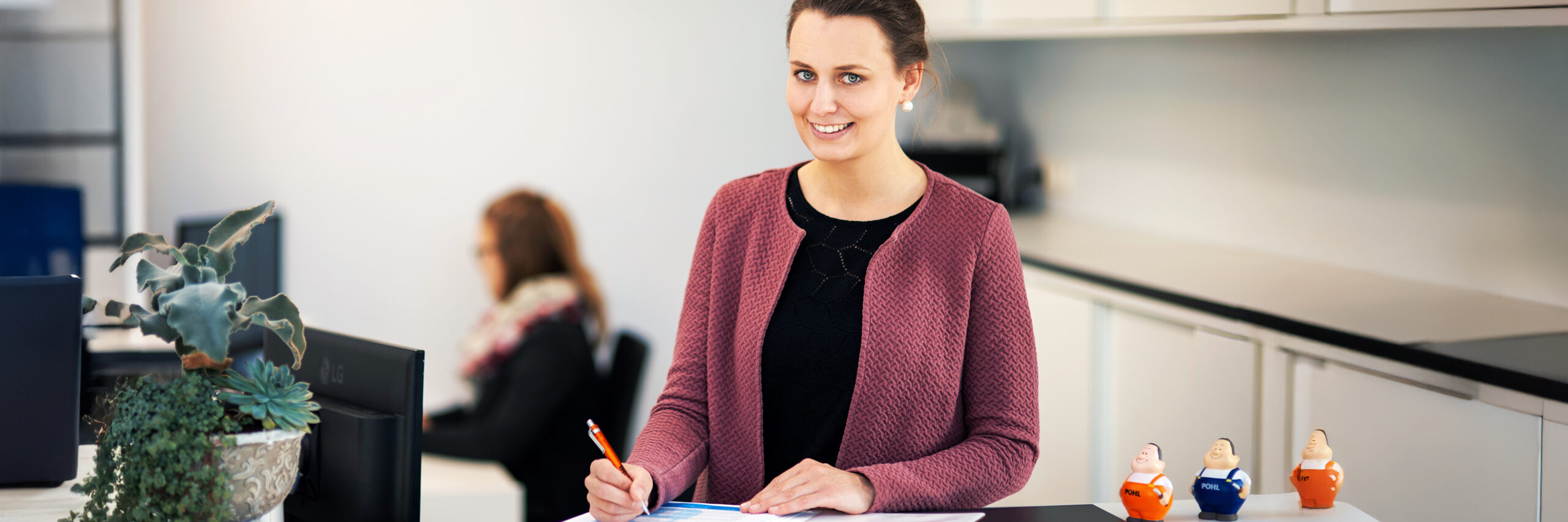In der Mitte füllt eine POHL-Mitarbeiterin an einem Tresen ein Formular aus. Links vorne steht eine Schale mit Pflanzen; rechts auf der Theke stehen zwei kleine Figuren mit POHL-Aufdruck. Im Hintergrund sitzt eine weitere Person am Schreibtisch.