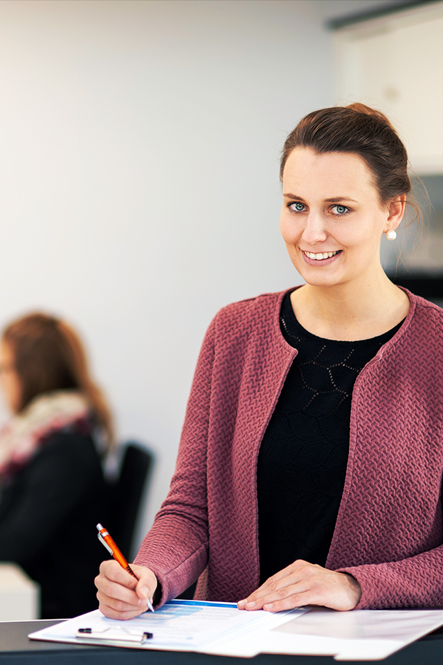 In der Mitte füllt eine POHL-Mitarbeiterin an einem Tresen ein Formular aus. Links vorne steht eine Schale mit Pflanzen; rechts auf der Theke stehen zwei kleine Figuren mit POHL-Aufdruck. Im Hintergrund sitzt eine weitere Person am Schreibtisch.