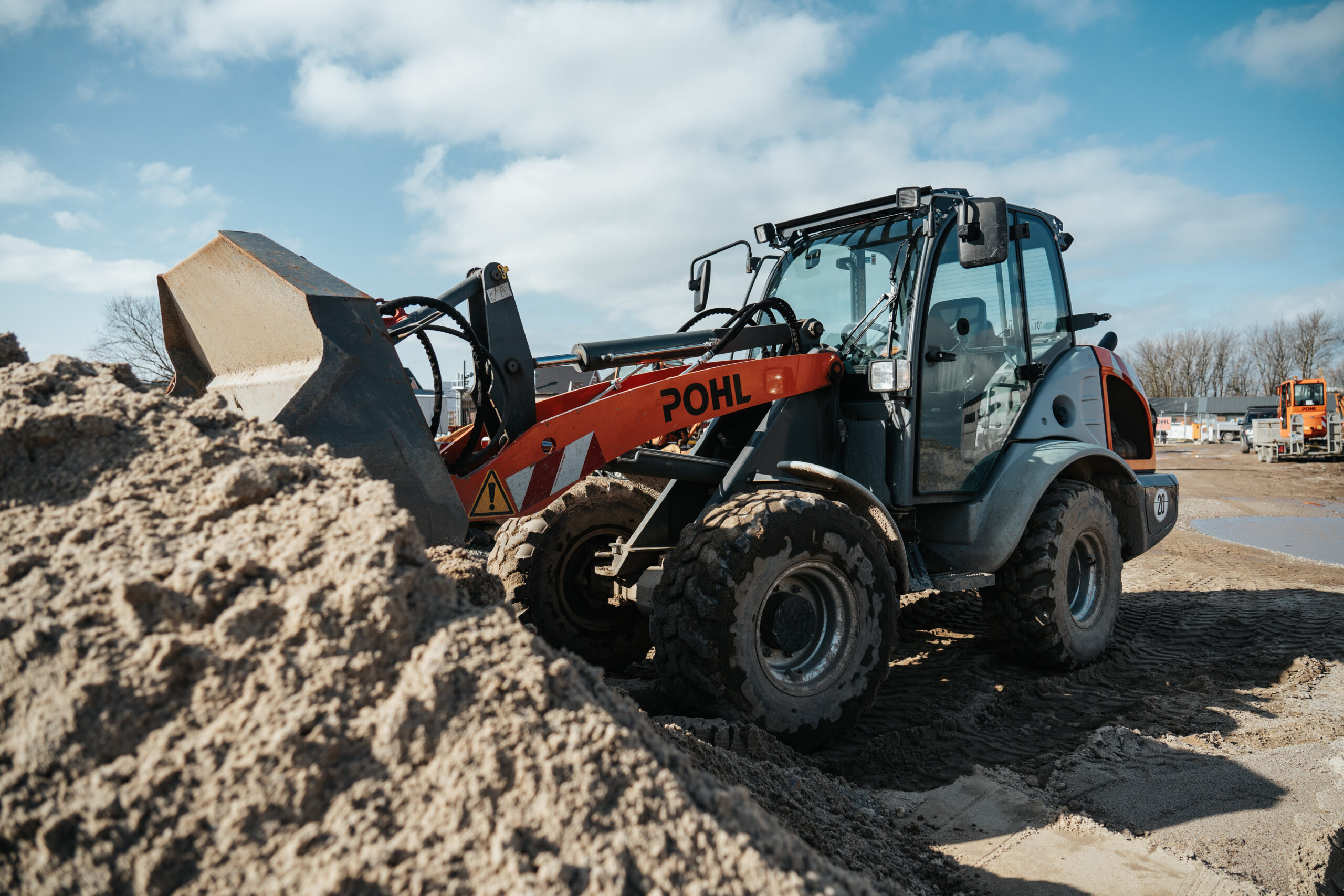 Standort Sylt: Vorne im Bild liegt ein hoher Sandhaufen. In der Mitte steht eine POHL-Maschine mit breiten Reifen und hochgehobener Schaufel. Rechts am Rand sind Fahrspuren im Sand. Im Hintergrund stehen weitere Fahrzeuge unter blauem Himmel.