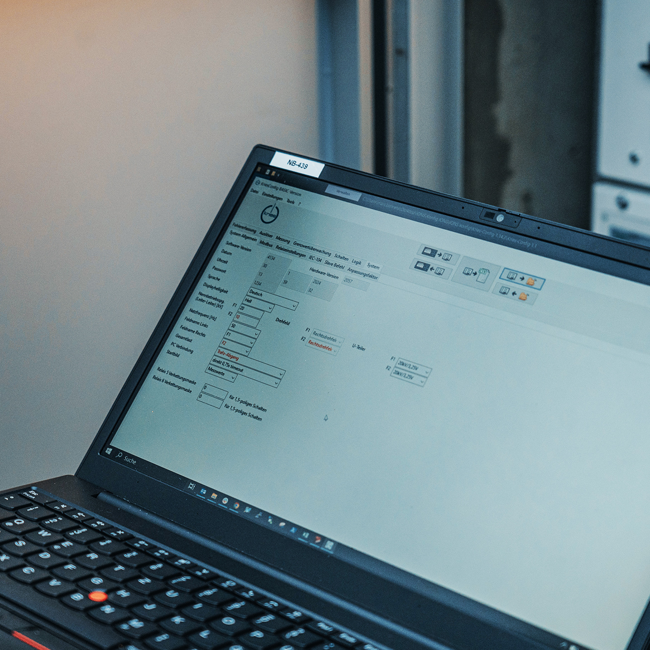 Vorne im Bild steht ein aufgeklappter Laptop; auf dem Bildschirm ist eine helle Konfigurationsoberfläche mit Eingabefeldern und Symbolen zu sehen. Rechts im Hintergrund stehen geöffnete Schaltschränke.