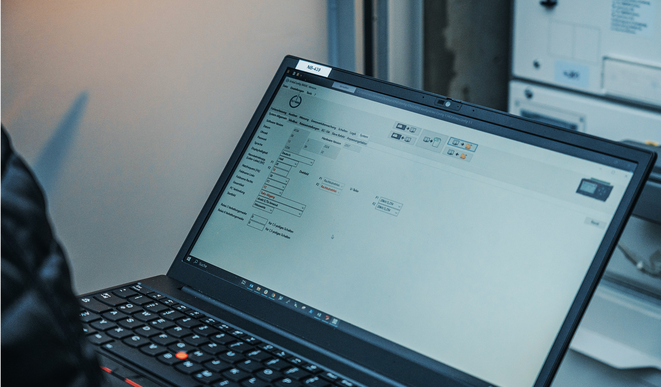 Vorne im Bild steht ein aufgeklappter Laptop; auf dem Bildschirm ist eine helle Konfigurationsoberfläche mit Eingabefeldern und Symbolen zu sehen. Rechts im Hintergrund stehen geöffnete Schaltschränke.