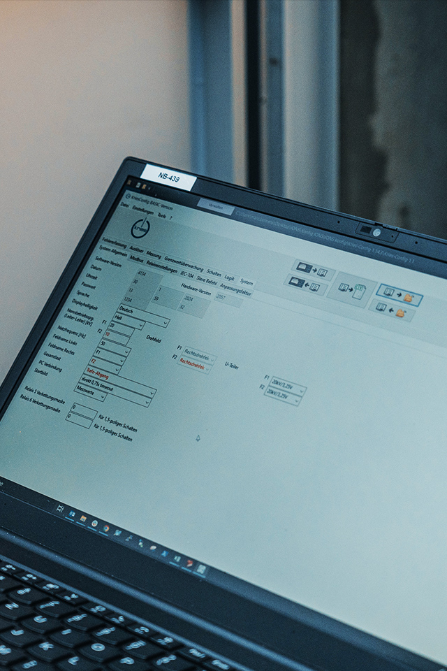 Vorne im Bild steht ein aufgeklappter Laptop; auf dem Bildschirm ist eine helle Konfigurationsoberfläche mit Eingabefeldern und Symbolen zu sehen. Rechts im Hintergrund stehen geöffnete Schaltschränke.