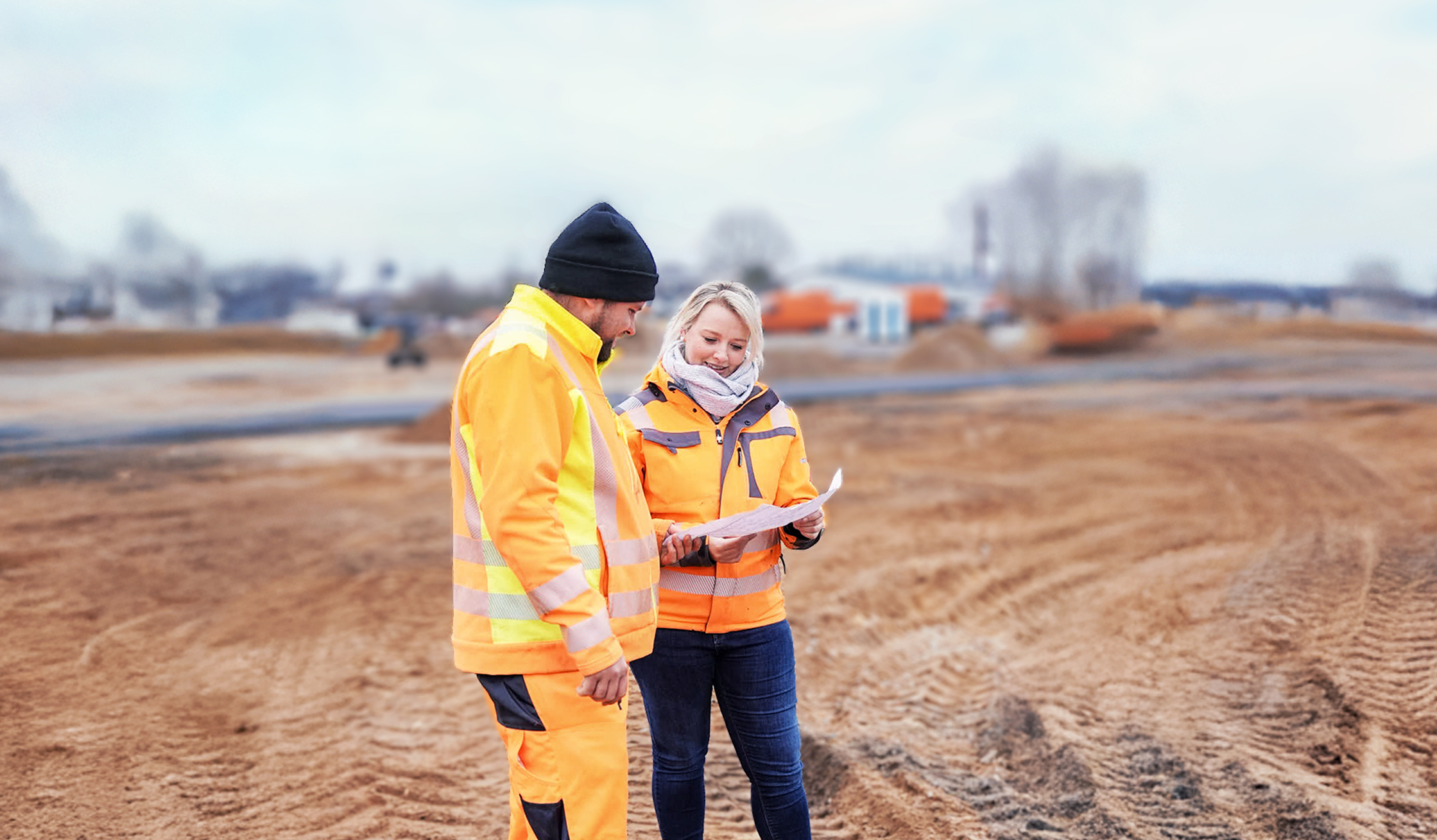 Weite, sandige Baustelle. Links im Vordergrund steht eine Person in gelber Warnjacke und Mütze; rechts daneben eine Person in orangefarbener Jacke mit Schal. Beide halten und betrachten einen Plan. Hintergrund unscharf mit Erdarbeiten und Fahrzeugen; kühles, trübes Tageslicht.