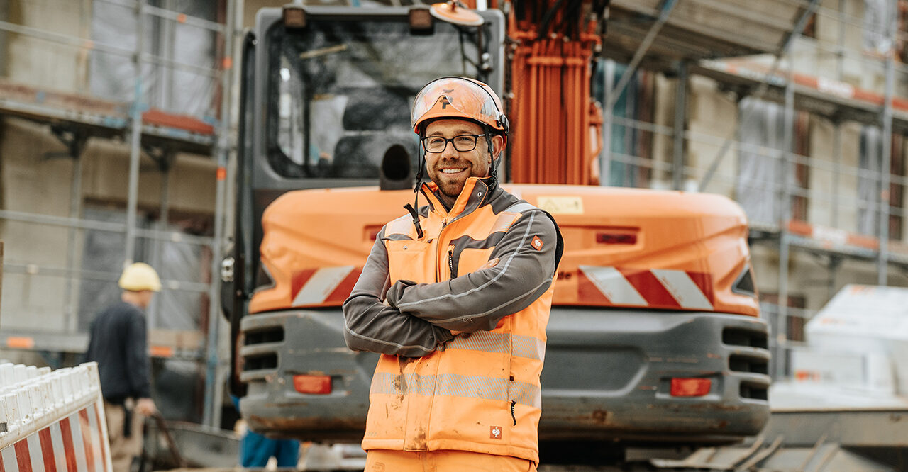 In der Mitte steht ein POHL-Mitarbeiter mit orangefarbenem Helm und Weste mit verschränkten Armen; direkt dahinter steht ein orangefarbener POHL-Bagger; im Hintergrund ist ein eingerüstetes Gebäude.