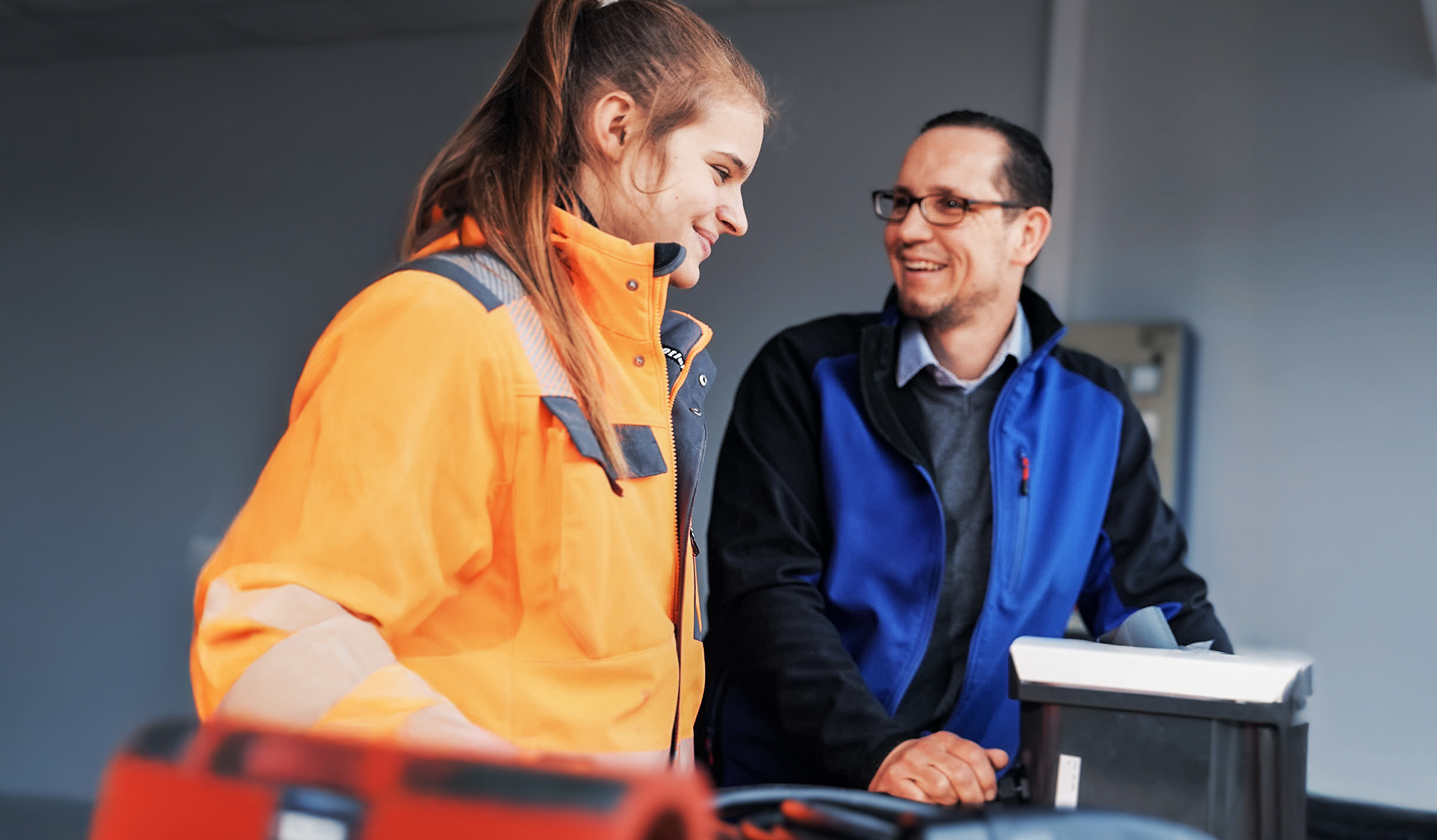 Innenraum mit grauer Rasterdecke. Links im Vordergrund beugt sich eine Person in orangefarbener Warnjacke über einen Werktisch. Rechts daneben steht eine Person in blau-grauer Arbeitsjacke, beide schauen sich an. Auf dem Tisch liegen ein Gerätegehäuse und Kabel; unscharfes Werkzeug vorne.