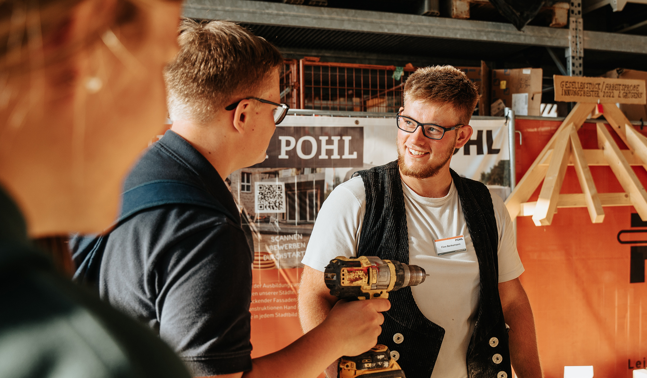 Innenaufnahme in einer Werkhalle, links vorn junger Mann mit Akkuschrauber spricht mit einem lächelnden Handwerker im schwarzen Zimmermanns-Gilet, beide vor einem orangefarbenen Stand mit QR-Plakat und Pohl Schriftzug.