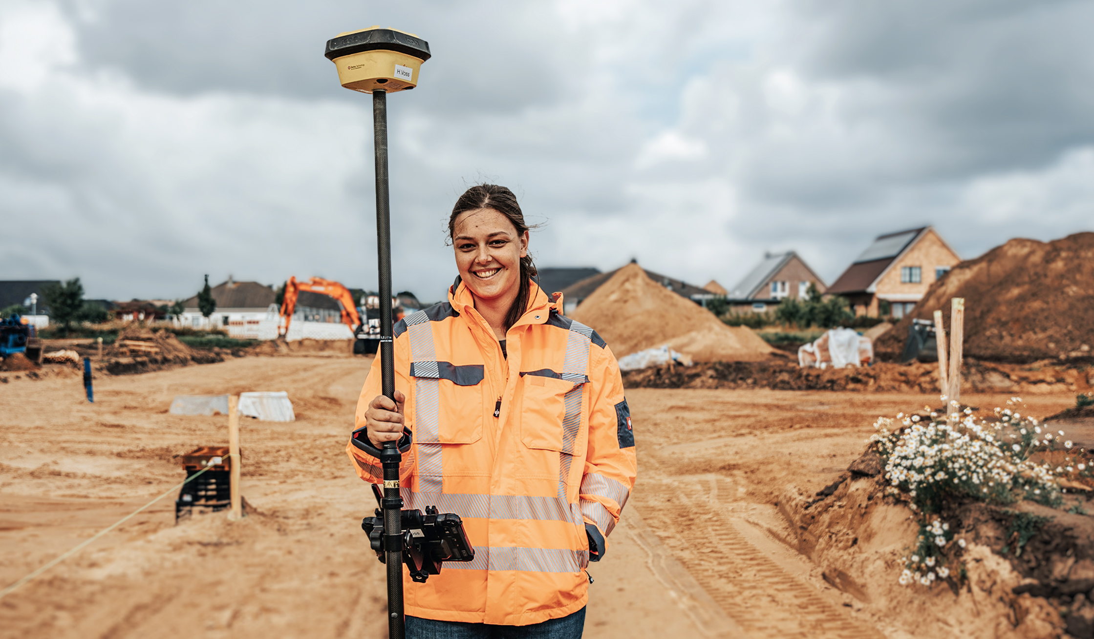 Zentriert im Vordergrund hält eine Frau in orangefarbener Warnjacke einen hoch aufgerichteten GPS/RTK-Vermessungsstab; sie schaut lächelnd in die Kamera. Links und rechts im Hintergrund eine sandige Baustelle mit Erdhaufen, Fahrspuren und einigen Häusern; rechts im Vordergrund weiße Wildblumen. Himmel bewölkt.