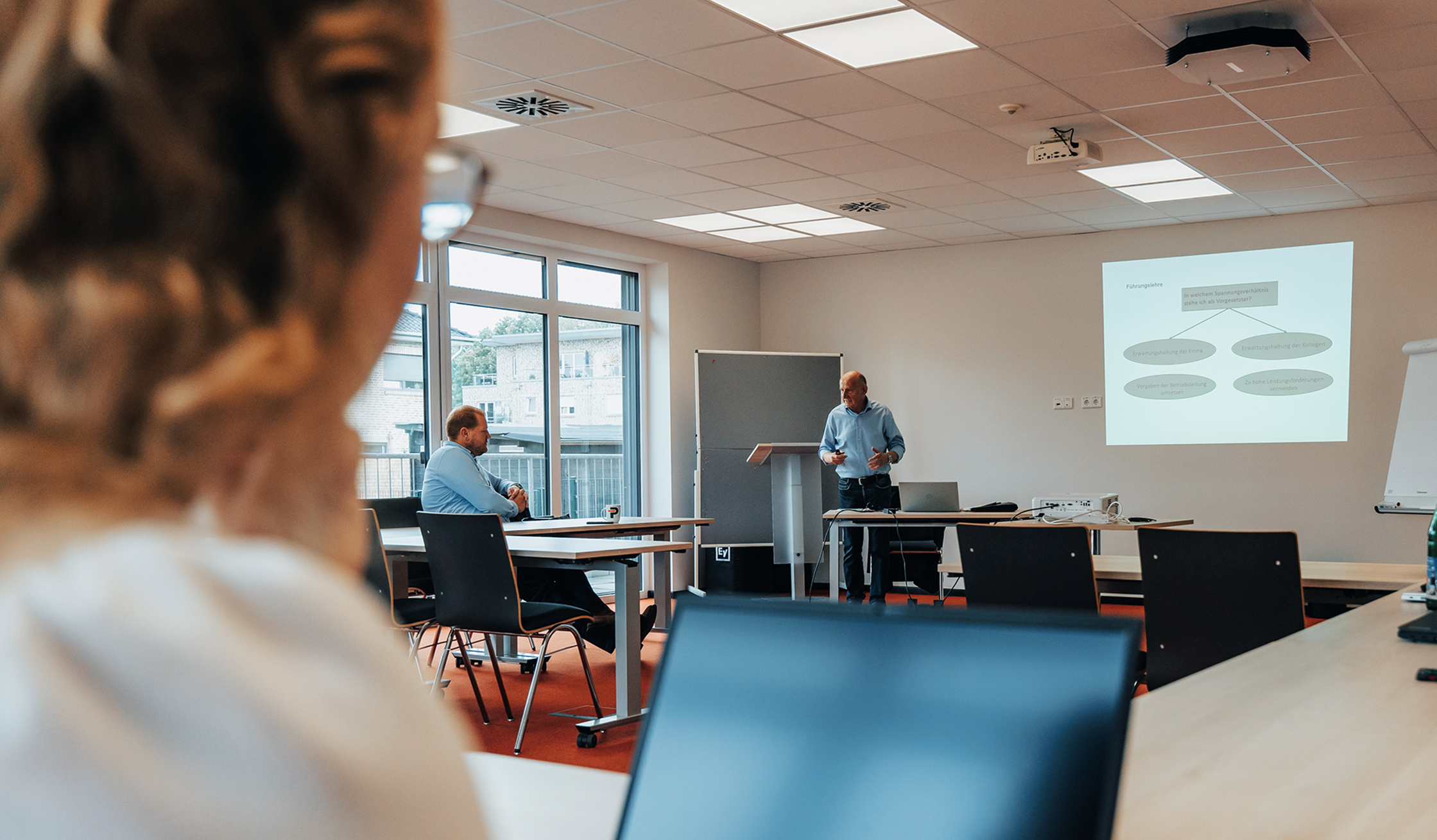 In der Mitte spricht ein Referent neben einer projizierten Folie; rechts steht ein Roll-Flipchart. Vorne links ist der Kopf einer Zuhörerin unscharf; links und hinten fällt Tageslicht durch große Fenster.