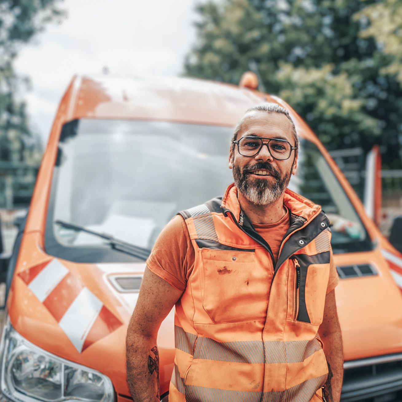 Halbporträt, Person leicht linksstehend vor orangefarbenem Transporter. Brille, grauer Bart, orange ärmellose Warnweste über T-Shirt, sichtbare Gebrauchsspuren. Hintergrund Straße und Fahrzeug unscharf, helles Tageslicht.