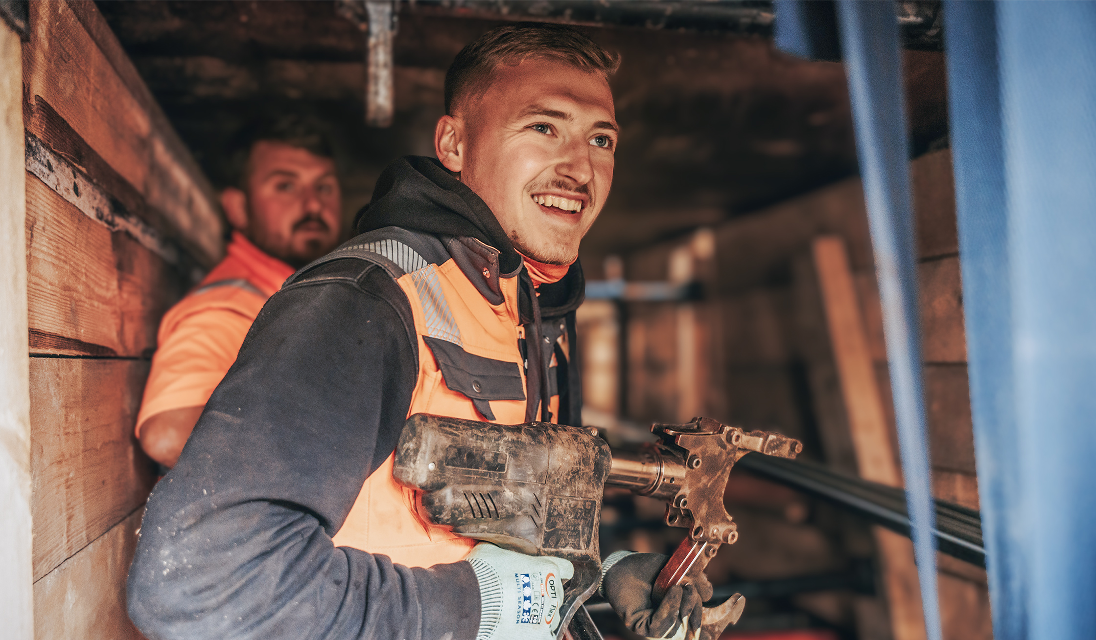 In der Mitte hält ein POHL-Mitarbeiter mit orange Weste und Handschuhen ein handgeführtes Werkzeug. Links hinten steht ein weiterer Mitarbeiter; die Wände bestehen aus dicht gesetzten Holzbohlen.