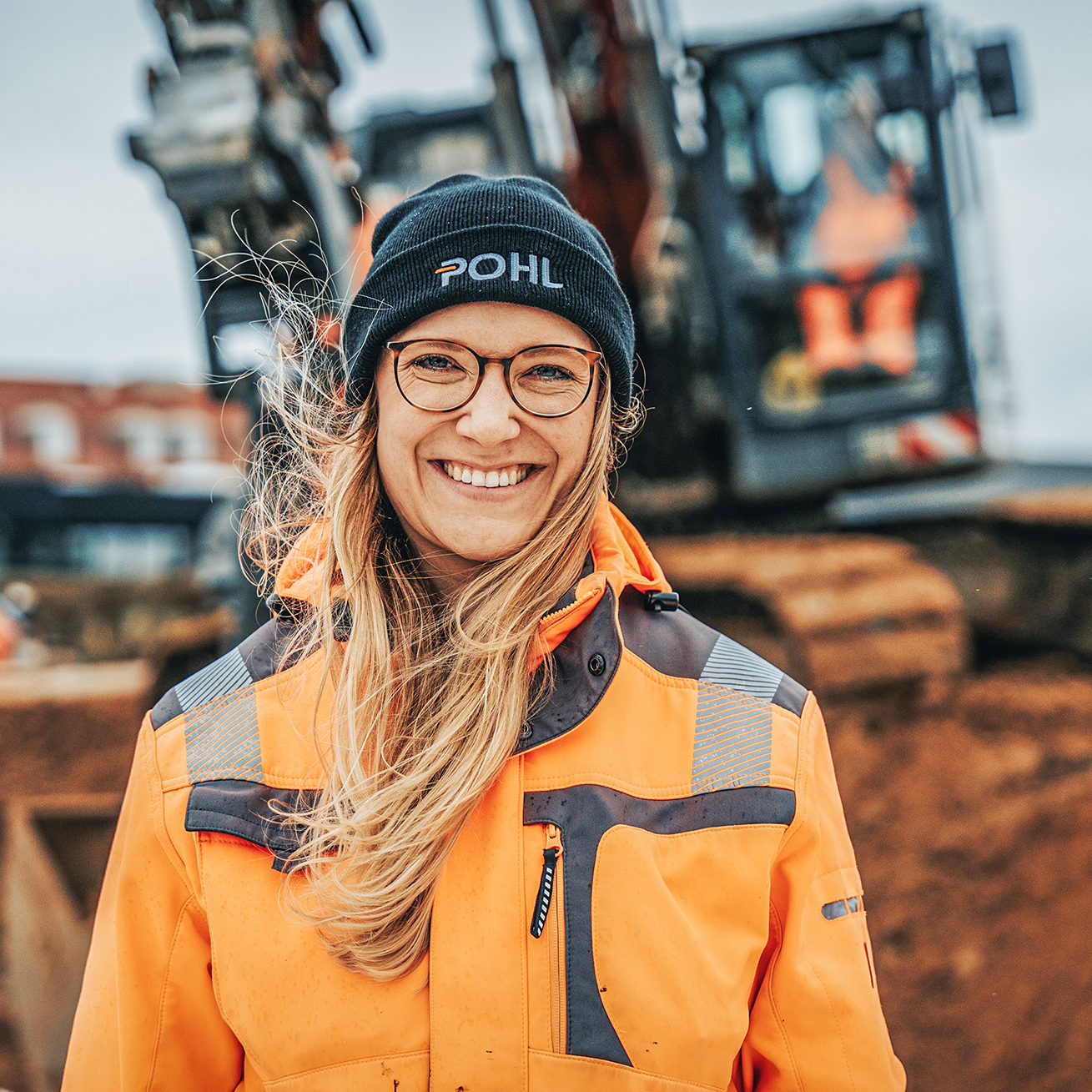 Kopf-Schulter-Porträt, Person mittig. Schwarze Mütze, runde Brille, orange Warnjacke; lächelnd, vom Wind bewegte Haare. Hintergrund unscharf: Baustelle mit Kettenfahrzeug und Erdhügeln, grauer Himmel.
