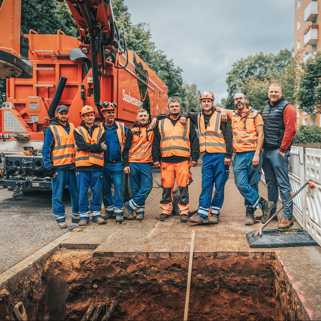 Vorne mittig steht eine Gruppe aus acht Personen in Warnwesten nebeneinander und blickt zur Kamera. Links hinten ein großer orangefarbener Saugbagger. Rechts entlang des Gehwegs weiße Absperrgitter. Im Vordergrund der Rand einer geöffneten Baugrube; hinten Wohnblöcke und Bäume.