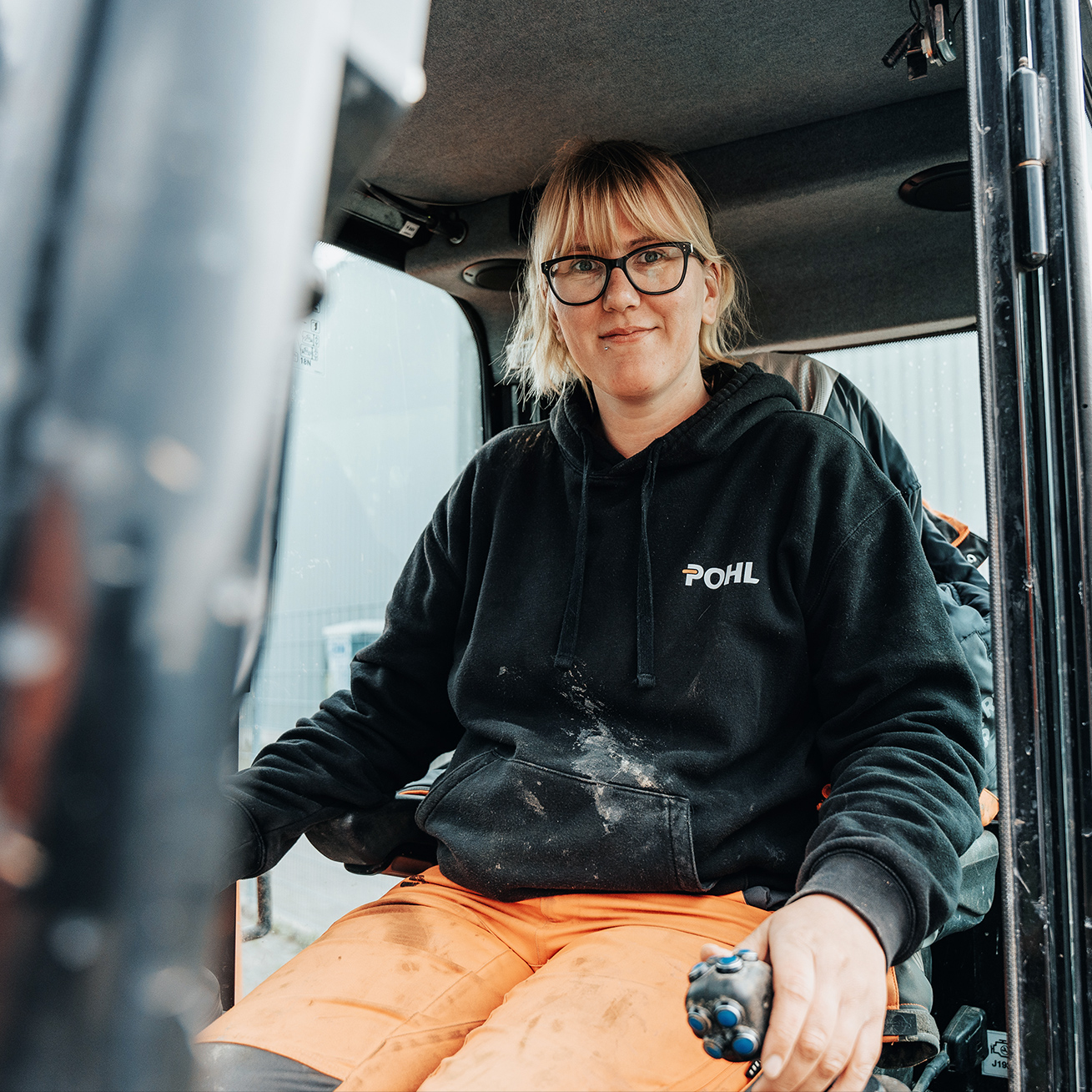 Vorne mittig sitzt eine Frau mit Brille im Führerhaus eines Minibaggers; schwarzer POHL-Hoodie, Hände an den Joysticks. Links begrenzt die geöffnete Kabinentür den Bildrand. Rechts und hinten ein Zaun und eine Hallenfassade auf dem Betriebshof; Kleidung sichtbar staubig.