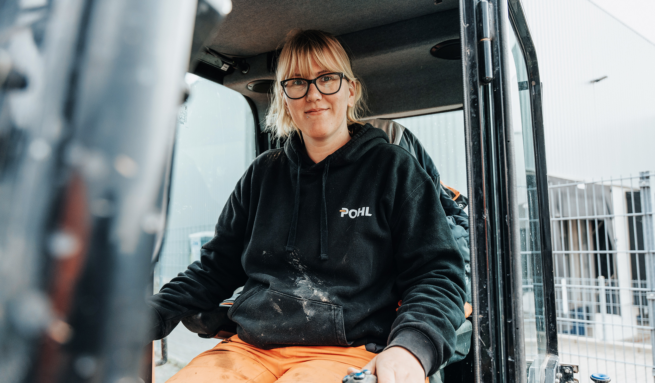 Vorne mittig sitzt eine Frau mit Brille im Führerhaus eines Minibaggers; schwarzer POHL-Hoodie, Hände an den Joysticks. Links begrenzt die geöffnete Kabinentür den Bildrand. Rechts und hinten ein Zaun und eine Hallenfassade auf dem Betriebshof; Kleidung sichtbar staubig.