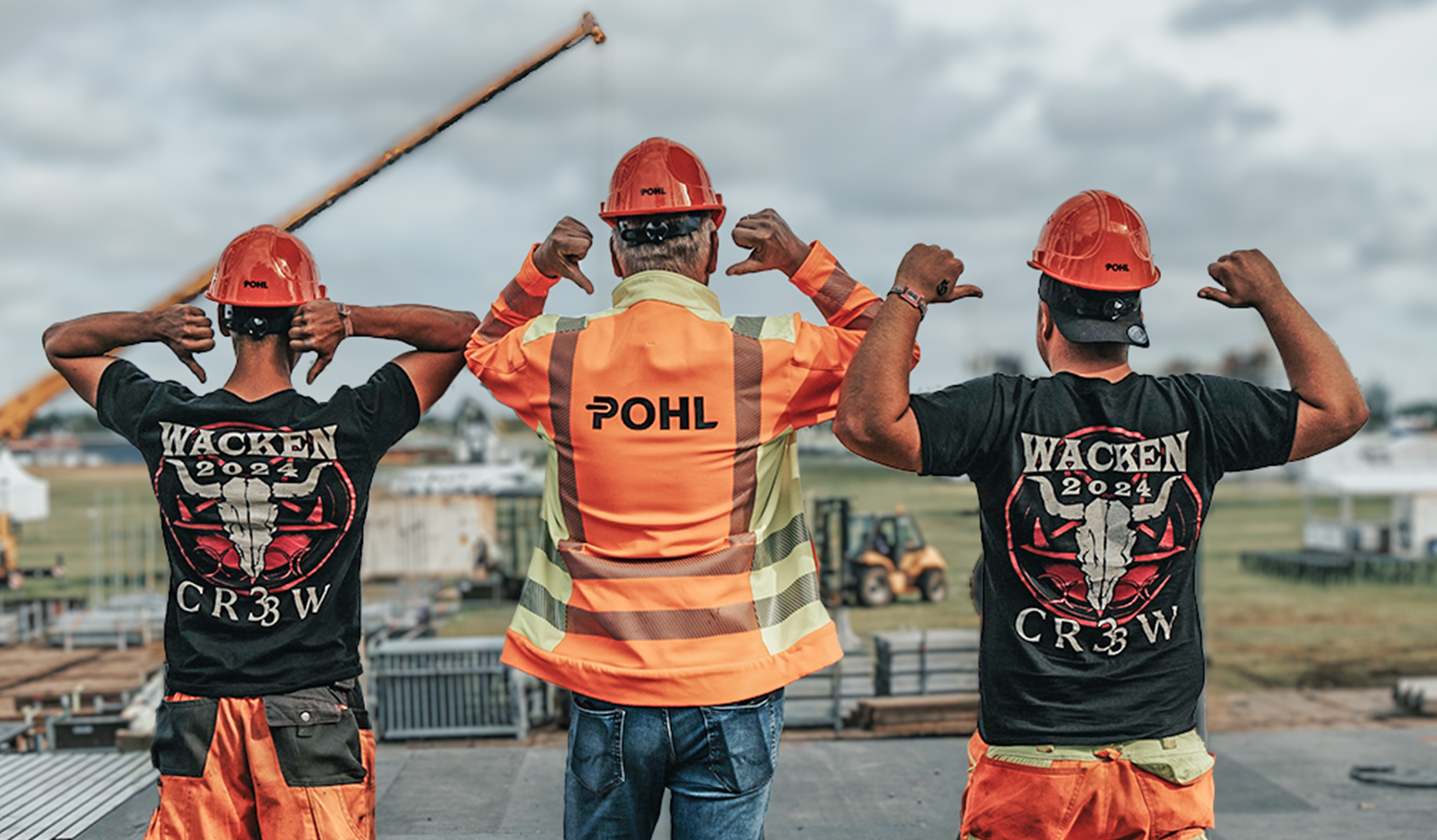 Drei Personen stehen auf einem Festivalgelände mit dem Rücken zur Kamera; die mittlere Person trägt eine POHL-Warnweste, die beiden äußeren „Wacken 2024 Crew“-Shirts.