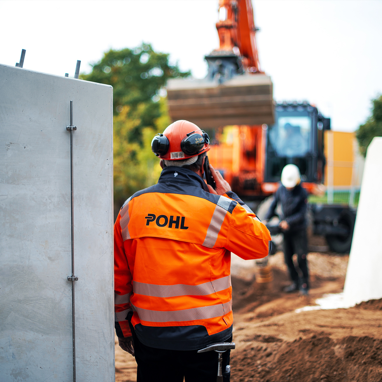 Mitarbeiter von POHL in orangener Schutzkleidung telefoniert auf einer Baustelle vor einem Bagger.