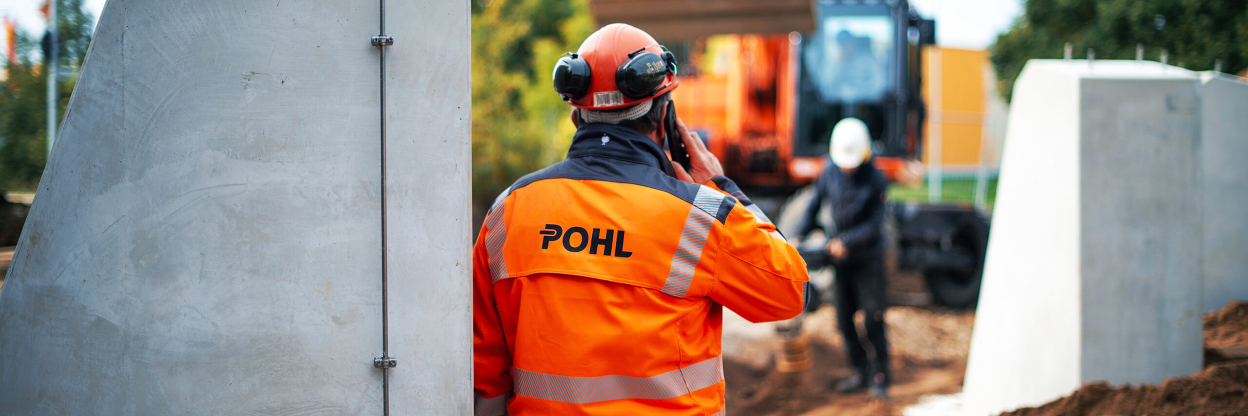 Mitarbeiter von POHL in orangener Schutzkleidung telefoniert auf einer Baustelle vor einem Bagger.