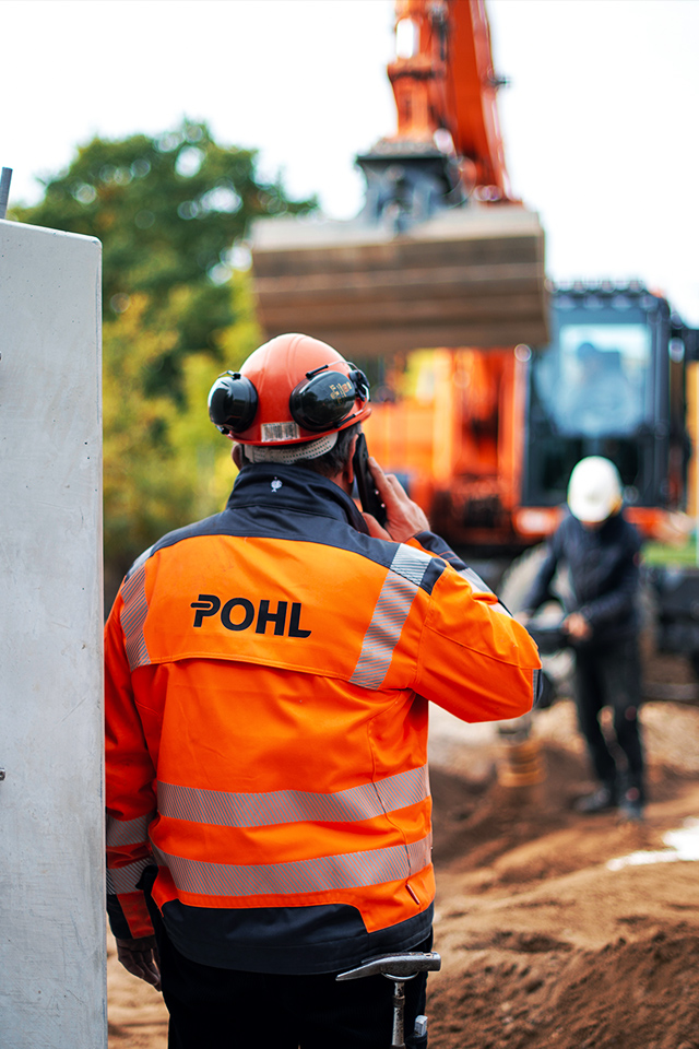 Mitarbeiter von POHL in orangener Schutzkleidung telefoniert auf einer Baustelle vor einem Bagger.
