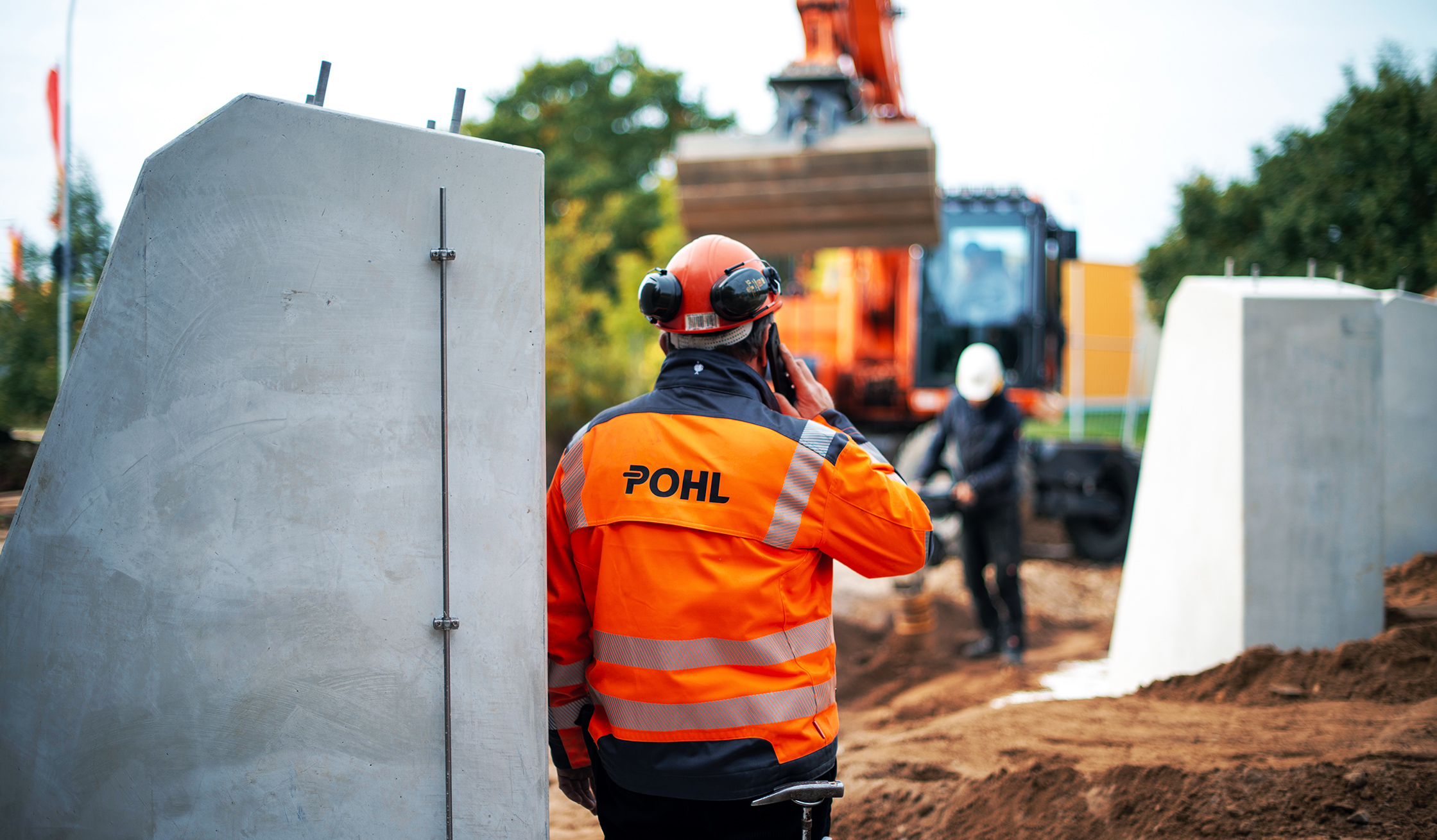 Mitarbeiter von POHL in orangener Schutzkleidung telefoniert auf einer Baustelle vor einem Bagger.