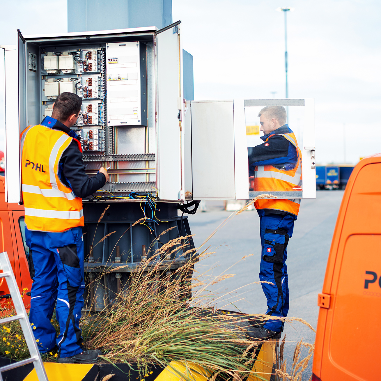 Zwei POHL-Elektroniker arbeiten an einem geöffneten Schaltschrank zwischen zwei orangefarbenen POHL-Service-Vans; Kabel und Klemmen sind sichtbar.