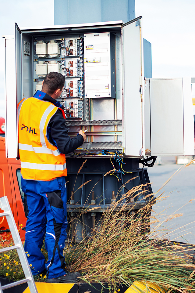 Zwei POHL-Elektroniker arbeiten an einem geöffneten Schaltschrank zwischen zwei orangefarbenen POHL-Service-Vans; Kabel und Klemmen sind sichtbar.