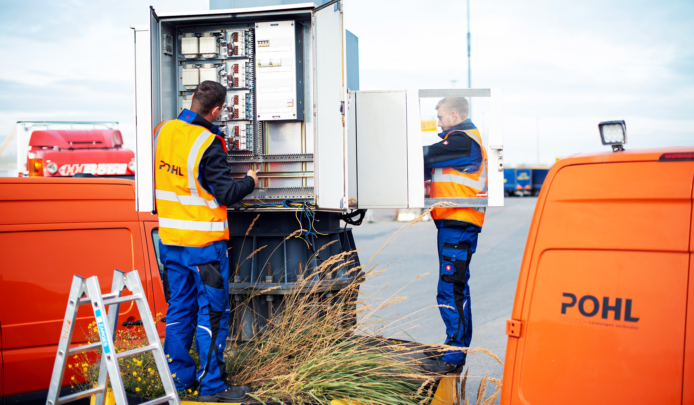 Zwei POHL-Elektroniker arbeiten an einem geöffneten Schaltschrank zwischen zwei orangefarbenen POHL-Service-Vans; Kabel und Klemmen sind sichtbar.