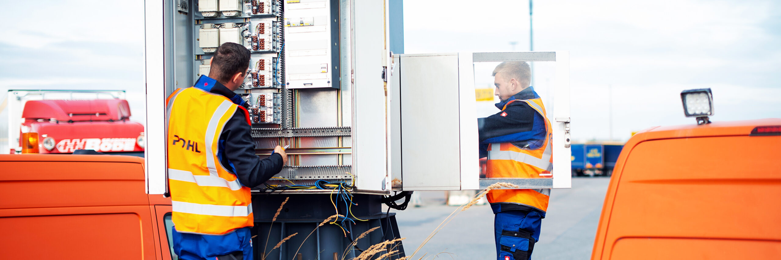 Zwei POHL-Elektroniker arbeiten an einem geöffneten Schaltschrank zwischen zwei orangefarbenen POHL-Service-Vans; Kabel und Klemmen sind sichtbar.