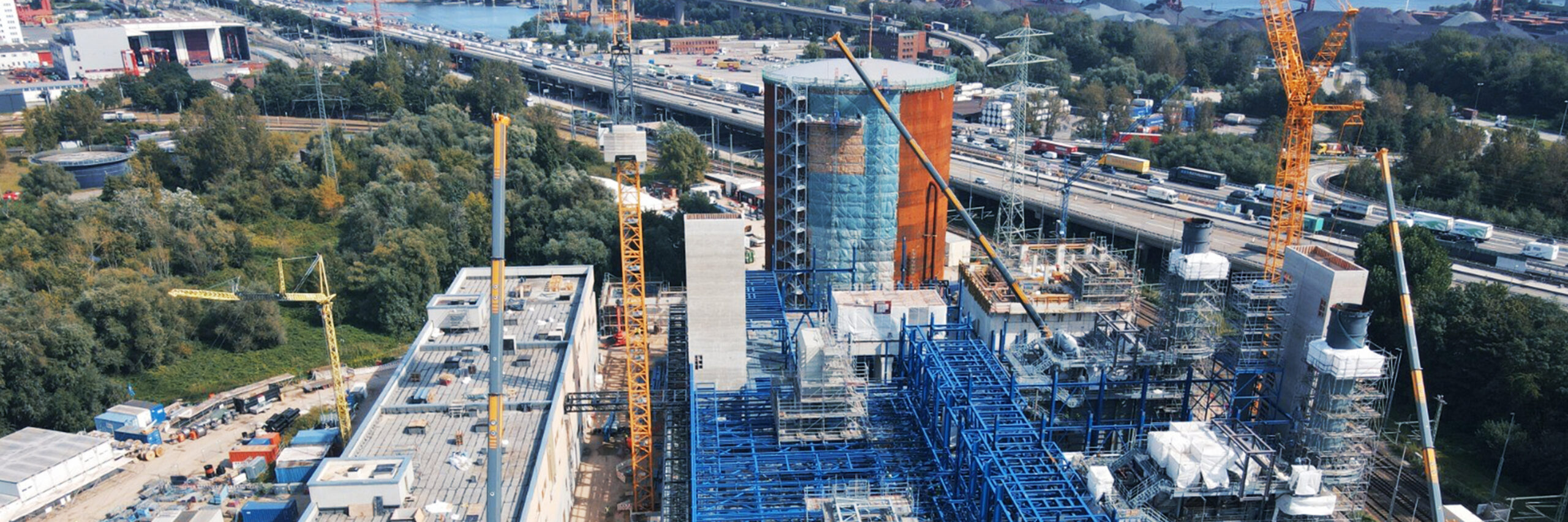 Referenz KWK Dradenau: Baustelle von oben: in der Mitte großes blaues Stahlgerüst, links und rechts Kräne, vorne Hallen, hinten Autobahn mit Autos.