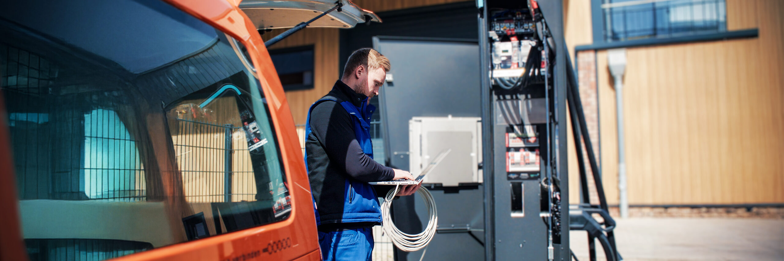 Ein POHL Mitarbeiter in blauer Arbeitskleidung arbeitet an einem offenen Schaltschrank. Er hält ein Kabelbündel und tippt auf einem Laptop, während ein orangefarbenes POHL-Fahrzeug hinter ihm steht.