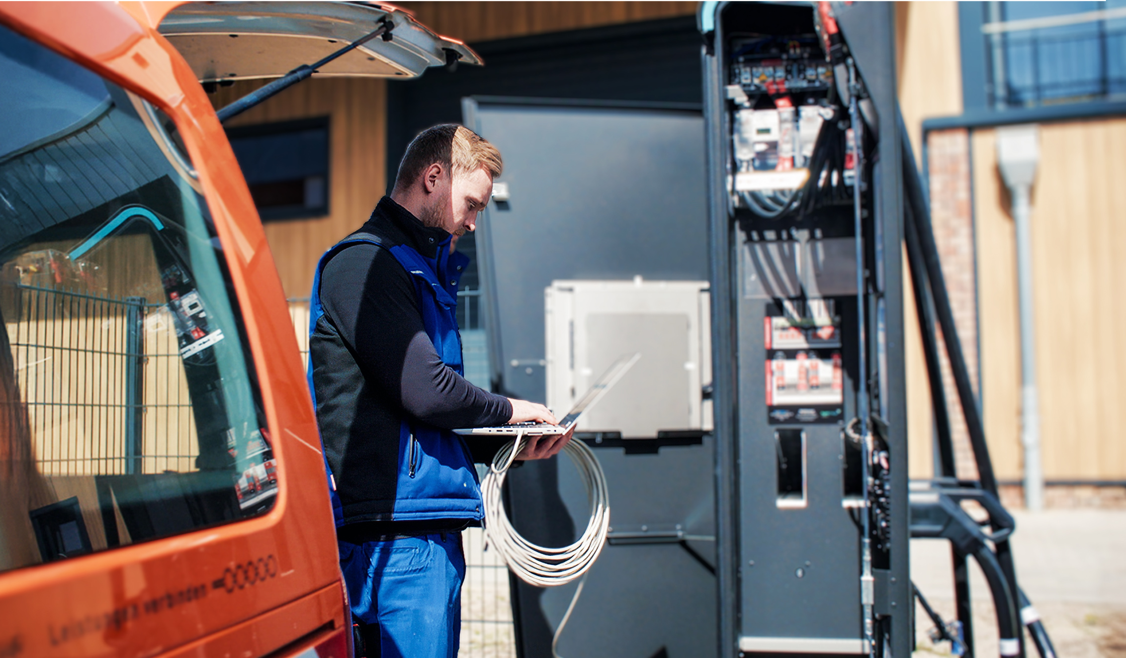 Ein POHL Mitarbeiter in blauer Arbeitskleidung arbeitet an einem offenen Schaltschrank. Er hält ein Kabelbündel und tippt auf einem Laptop, während ein orangefarbenes POHL-Fahrzeug hinter ihm steht.