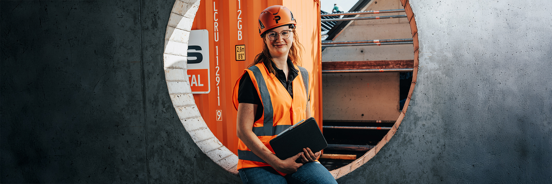 In der Mitte sitzt eine POHL-Mitarbeiterin mit orangefarbenem Helm und orangefarbener Weste in einer runden Öffnung einer grauen Betonwand und hält ein Tablet. Rechts im Bild stehen Gerüste und Bauteile. Links im Bild ist ein orangefarbener Container mit weißem Schild zu sehen. Im Hintergrund öffnet sich die Baustelle hinter der Wand.