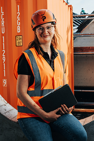 In der Mitte sitzt eine POHL-Mitarbeiterin mit orangefarbenem Helm und orangefarbener Weste in einer runden Öffnung einer grauen Betonwand und hält ein Tablet. Rechts im Bild stehen Gerüste und Bauteile. Links im Bild ist ein orangefarbener Container mit weißem Schild zu sehen. Im Hintergrund öffnet sich die Baustelle hinter der Wand.