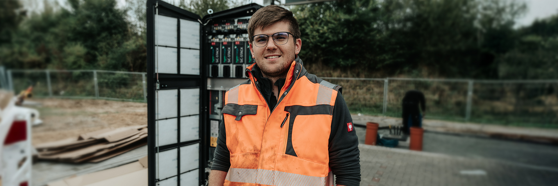 In der Mitte steht ein POHL-Mitarbeiter mit orangefarbener Warnweste vor einem geöffneten Technikschrank; links im Bild sind Kabel und Bauteile sichtbar, im Hintergrund steht ein Bauzaun vor Bäumen.