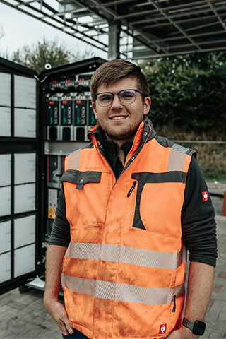 In der Mitte steht ein POHL-Mitarbeiter mit orangefarbener Warnweste vor einem geöffneten Technikschrank; links im Bild sind Kabel und Bauteile sichtbar, im Hintergrund steht ein Bauzaun vor Bäumen.