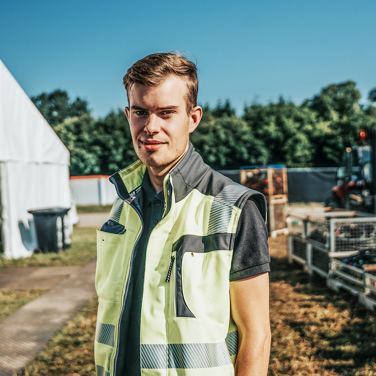 Auf einem Eventgelände steht ein POHL-Mitarbeiter in heller Warnweste vor einem weißen Zelt und Materialkisten; Hintergrund unscharf.