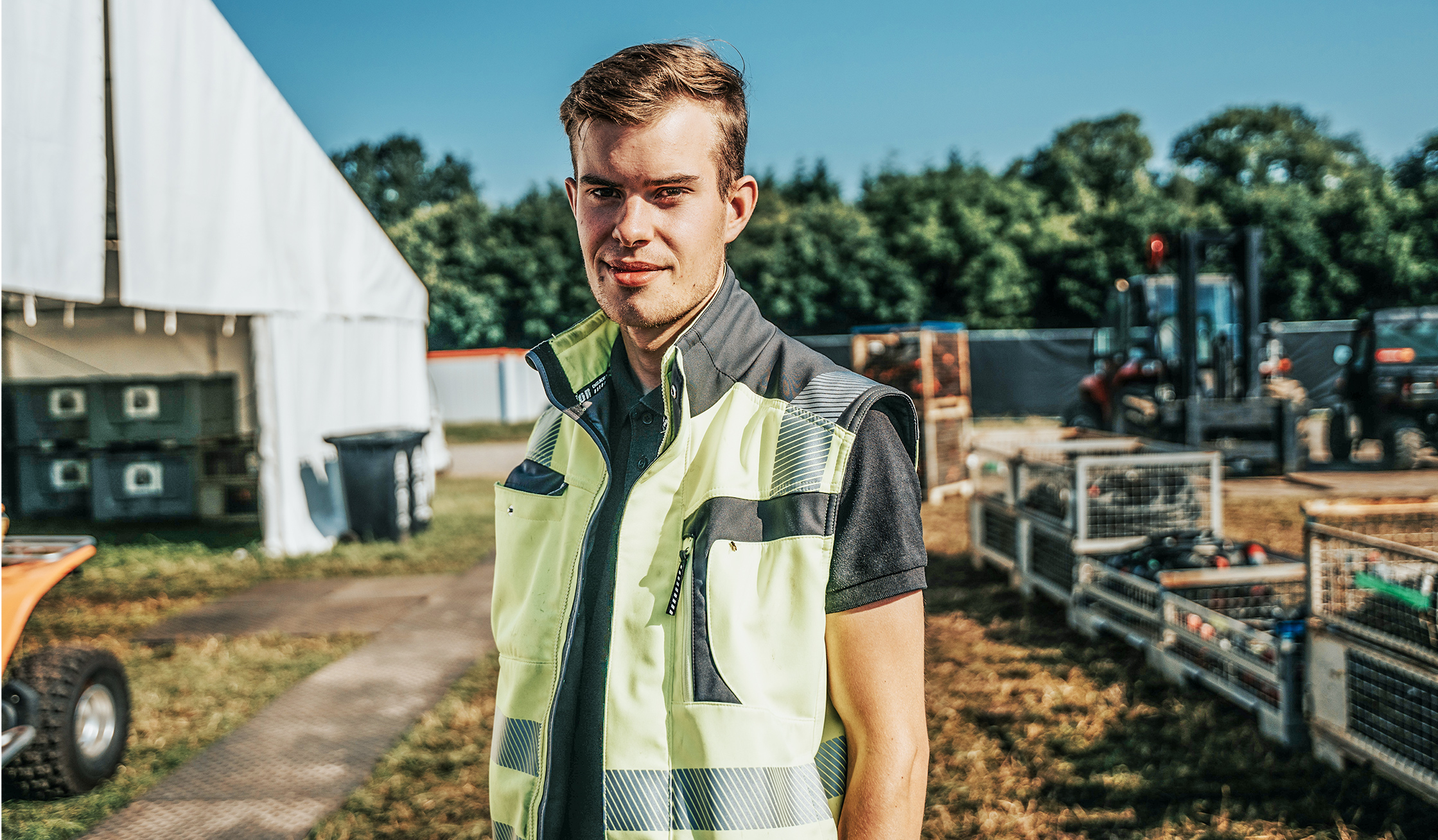 Auf einem Eventgelände steht ein POHL-Mitarbeiter in heller Warnweste vor einem weißen Zelt und Materialkisten; Hintergrund unscharf.